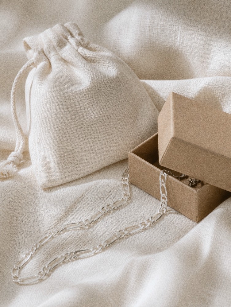 A fine sterling silver Figaro chain resting on soft ivory linen beside a linen drawstring pouch and an open kraft gift box