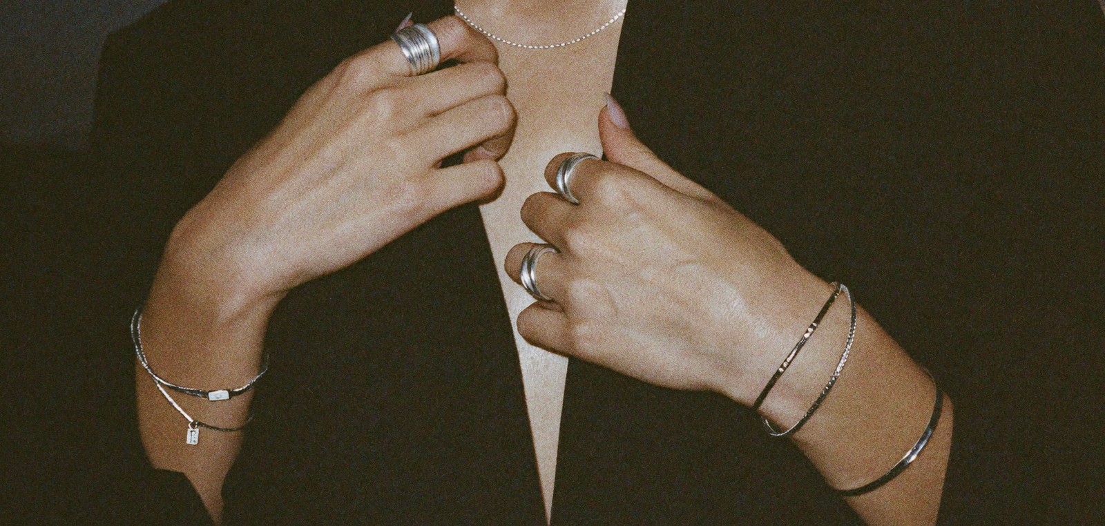 Two hands stacked with sterling silver rings, cuffs and a fine chain bracelet, fastening a necklace in warm low light