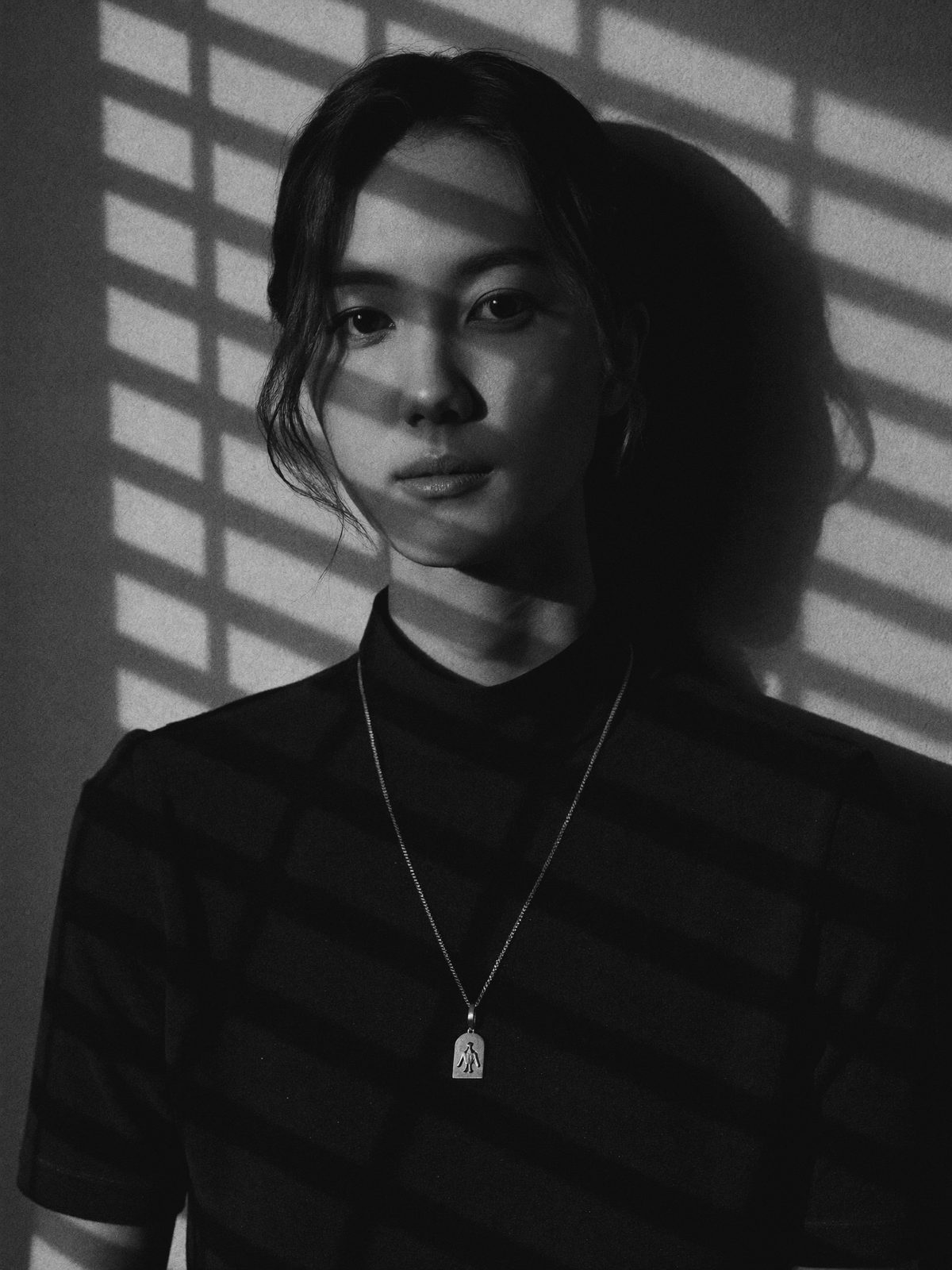 A woman lit by venetian-blind light in the dark, a fine sterling silver pendant catching a single bar of light — shot on black-and-white film